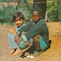 Two poor boys sit on dirt outdoors. One is smiling, barefoot, with shaggy dark hair. The other with a serious look, wearing a dark sweater, shorts, and worn shoes. A barbed wire fence runs across the background.