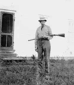 picture of a man with a moustache, standing, holding a rifle and wearing a hat