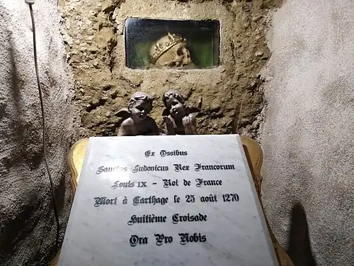A human skull with a crown in a niche. Outside of the niche, two angel putti hold a marble plate inscribed 'Ex Ossibus / Sanctus Ludovicus Rex Francorum / Louis XI - Roi de France / Mort à Carthage le 25 août 1270 / Huitième Croisade / Ora Pro Nobis'.