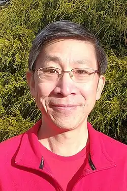 Headshot of an Asian-American man with short dark hair and glasses, wearing a red shirt and jacket, standing in front of a chartreuse evergreen bush