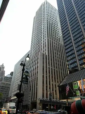 The Daily News Building's facade as viewed from 42nd Street, looking southeast. The building is a rectangular tower with a white-brick facade, interspersed with vertical bands of windows and dark-colored spandrel panels. There are several setbacks on the facade. A black glass tower is visible on the right.