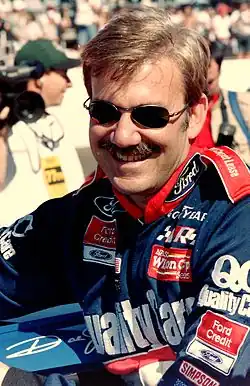A mustached man in his early forties wearing sunglasses and blue and red racing overalls