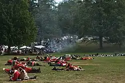 An open grassy area is strewn with bodies, most of them in red and white uniforms, although some with blue coats are visible farther back. In the distance there are white tents and a crowd of people, and a small thin cloud of smoke obscures the view a little.