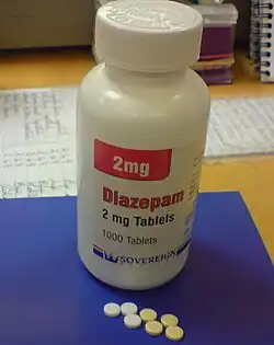 White bottle with red and black labels on a blue pad atop a desk. Also on the pad are seven small pills.