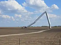 A collapsed wind turbine over open country, bent sharply below its midpoint