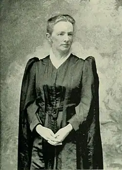 A white woman, standing, in a black clergy robe, with hands folded.