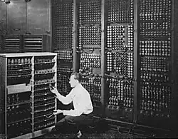 Black-and-white image of a man replacing one vacuum tube out of hundreds in early computer