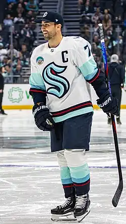 Ice hockey player holding a stick and wearing a cap while in a blue Seattle Kraken jersey, featuring a letter A on the top right