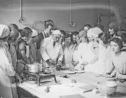 Middle-aged white man with large moustache, white coat and skull-cap, surrounded by young men and women, at a cookery workbench