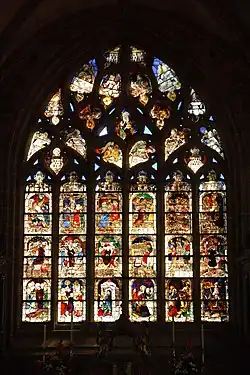 The chevet window with scenes from the Passion (Christianity). Scenes in the top row depict the raising of Lazarus, the entry into Jerusalem, the last supper and Joan of Arc on horseback. In the next row is a scene depicting Jesus in the garden of olives, then his arrest and his being ridiculed, his appearance before Herod, the flagellation with Jesus tied to a tree, the carrying of the cross. In the lower row are two scenes involving the crucifixion, then Jesus being brought down from the cross and the preparation for his burial (the "Mise au Tombeau" followed by Jesus resurrected and emerging from the tomb and then his descent into limbo to save some souls trapped there