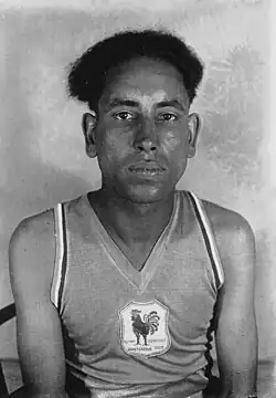 A black and white portrait photograph of a man with short and frizzy black hair. He is wearing a jersey with an image of a rooster on it.