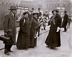Emmeline Pethick Lawrence receiving a bouquet of flowers from Jennie Baines, Flora Drummond and Frederick Pethick Lawrence watching.