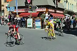 A line of road racing cyclists, including one in a red, white, and black jersey riding directly in front of one with a prominently yellow jersey. Spectators watch from the roadside.