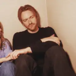 A sullen bearded ginger man holds someone's hand off-frame as he sits in the corner of a room.