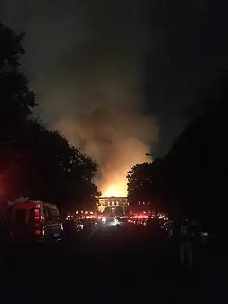 Photograph of the museum fire lighting the night sky at the end of a long, dark street lined with vehicles at either side