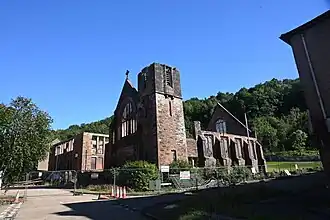 Part demolished roofless fenced off stone building with tower and butressed side walls, against a wooded hillside background on a sunny day with a deep blue sky