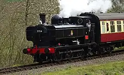 A green pannier tank locomotive is seen from above and to the front as it waits at a station with two railway tracks and platform. The platform on the left includes a small wooden shelter, benches, lamps, flowers, and, near the end of the platform, three milk churns positioning across the platform to prevent people passing. The platform on the right, where the train is waiting, includes a brick building, more flowers, and again, three milk churns arranged to stop people passing. There is also a signal near the locomotive, and its black and white semaphore arm is pointing downward. The train includes seven coaches in maroon. A plume of grey smoke comes from the locomotive chimney, and a smaller plume of white steam from the safety valve near the cab.