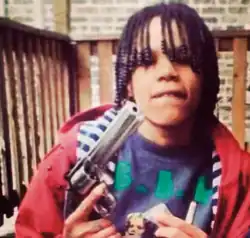 An image of Gakirah Barnes. She is holding a revolver in her hand while wearing a red hoodie with a striped interior. She is looking slightly away from the camera.