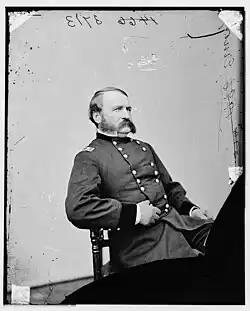 Black and white photo of a seated man with a large moustache and muttonchops. He wears a dark military uniform with two rows of buttons, and the two stars of a major general on the shoulder tabs.