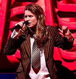 A woman with long brown hair holds a microphone close to her mouth while wearing a pinstripe suit and a polka dot tie