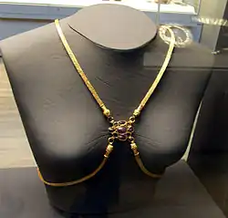 Frontal view of a grey female bust with four gold bands of many fine links, two draped over the shoulders and two plunging from the breast bone sweeping below the breasts and going behind the back. The four converge between the breasts, where each band ends in a head which connects to a centerpiece, a purple gem surrounded by eight smaller sockets, four empty and four with red stones.