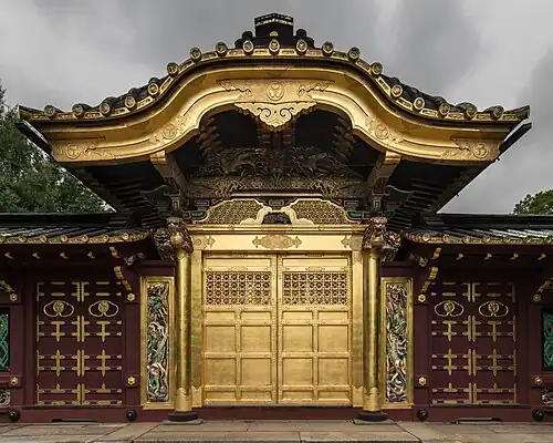 The front of a Shinto Shrine, colored gold and brown, with a golden overhang to the front doors. There are carvings both above and to the sides of the main doors. The sky is overcast and dark.