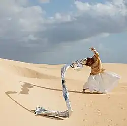 A red headed woman wearing a beige shirt and white skirt stands in the middle of a desert with the wind blowing her arms and a large drapery that flows from her hand.