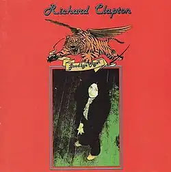 Background is bright red. At top is the artist's name in stylised, blue script. Below is a depiction of a flying tiger, in orange with black stripes. Its jaws open and claws are reaching forward. Just below in smaller script is the album's name on a yellow ribbon. At the bottom two-thirds is an overhead shot of the artist. He wears a dark top and dark pants; he has a multi-coloured shirt visible. He stands on a dark green-black wooden floor with his hands folded in front of him.