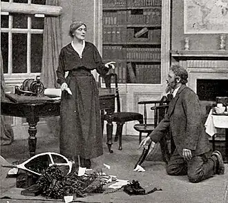 Standing woman in drawing room with bearded man kneeling amid debris