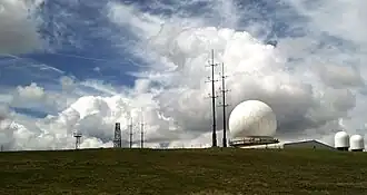 Photo shows a large white golfball-style radar dome, with two smaller radar domes and a number of other antenna; all set against a dramatic cloudy sky.
