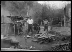 black-and-white photo of Māori man with male teenager and children in rustic cooking area