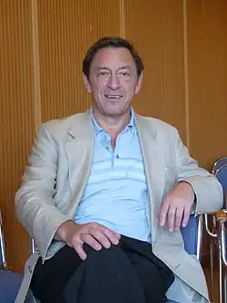 Guy Standing is sitting on a blue chair in front of a wooden wall. His right hand is on his knee and his left arm rests on the back of the chair.