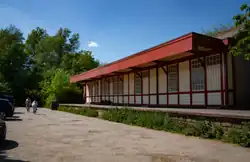 View of the disused but maintained station building at Halton from the former trackbed, parked cars on left, station on right, two cyclists in middle. Building painted in cream and red