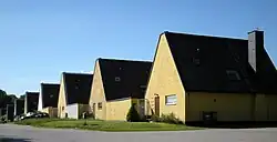 Image shows a row of yellow houses with dark roofs. Lawns have green grass, and the sky is blue.