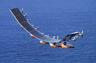 Helios Prototype in flight. The craft consists of a curved series of connected solar panels that have propellers behind them and fins on the bottom.
