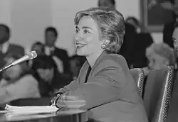 Photograph of Clinton making a presentation sitting at a table in front of a microphone
