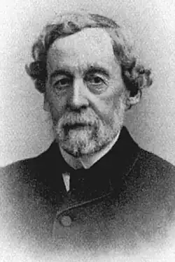 An older man in a thick suit coat with unkempt curly hair and a cropped white beard