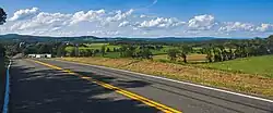 A rural landscape with a mix of fields and woodlots and low wooded mountains extends in the distance beneath a blue sky full of clouds. From bottom right to center left a paved road with a yellow stripe in the middle fills the image.