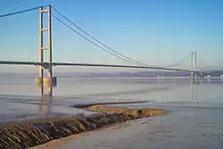 The near pier of a suspension bridge spanning calm blue waters of a wide river estuary.