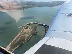 A view of Hurst Point and its lighthouse from the cockpit of a small aircraft. The lighthouse is displayed in the middle of the image with the aircraft wing to the right. In the background is the Isle of Wight