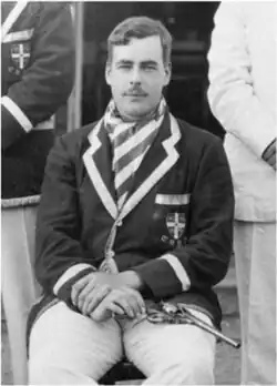 Black-and-white photograph of a young moustachioed man in a purple blazer, with a revolver on his lap.