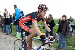 A road racing cyclist wearing a black and red jersey with white trim riding down the road. Spectators are visible on the roadside.