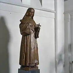 Wooden carving of a full length nun standing holding a large quill pen in her right hand and a crucifix in her left