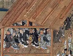 In a wooden building open at the sides, a troop of monks in black or grey robes dance in a circle.