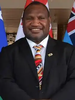James Marape in a suit in front of a Solomon Islands flag, Tongan flag, and Tuvaluan flag