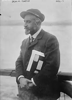 An older bearded man with a hat wearing a tie and coat. He is keeping a pile of papers or documents under his arm.