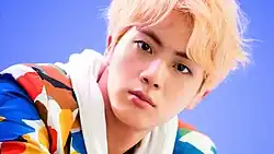 A headshot of Jin a colorful suit and blonde hair