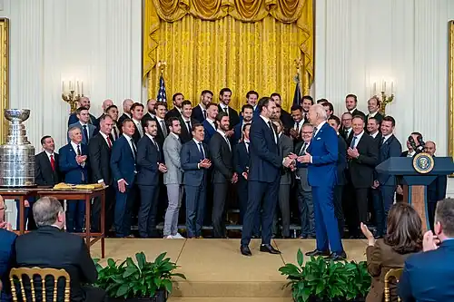 Two men in suits shake hands on stage with three rows of men observing behind them. The Stanley Cup sits on the left of the rows.