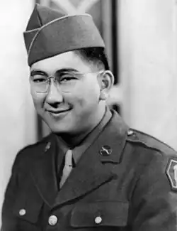 Head and shoulders of a smiling young man with dimples and round wire-framed glasses wearing a garrison cap and a military jacket over a shirt and tie