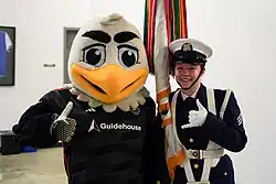 A black and white costumed bald eagle mascot with exaggerated features and an orange beak raising his wings. He wears a black soccer jersey with a white Volkswagen logo and the team's shield on it.